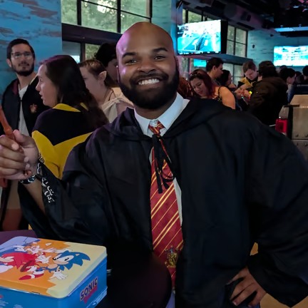Beaming wizard in robe and house tie shows off his wand at a Wizards Night Out bar.