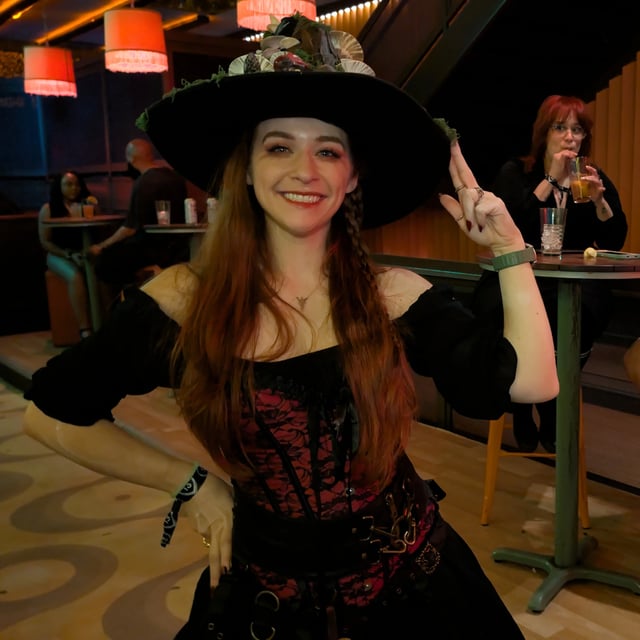 Wand-toting witch in floral pointed hat and corset gown grins inside a softly lit wizard-crawl lounge.