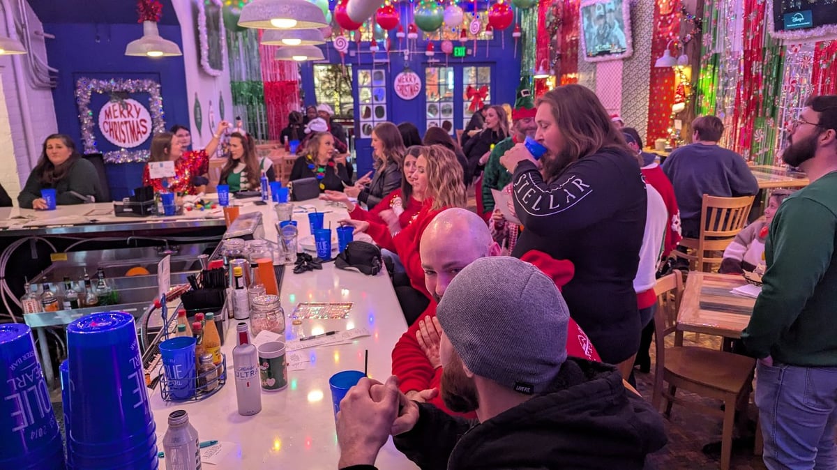 Crawlers in sweaters and beanies crowd a bar under a Merry Christmas wreath and tinsel curtains