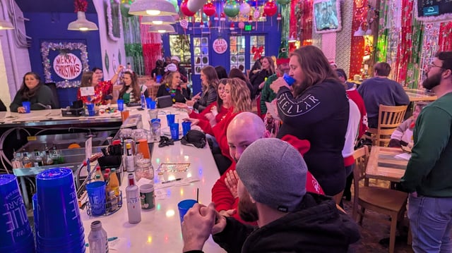 Crawlers in sweaters and beanies crowd a bar under a Merry Christmas wreath and tinsel curtains