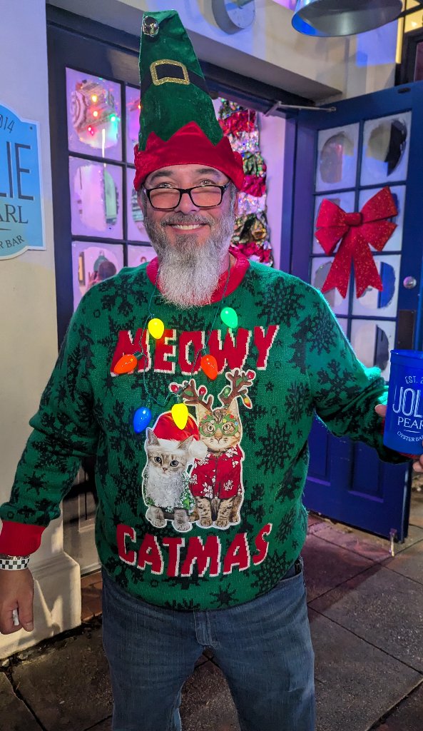 Bearded guy beams in a velvet elf hat and a Meowy Catmas sweater outside the Jolie Pearl bar