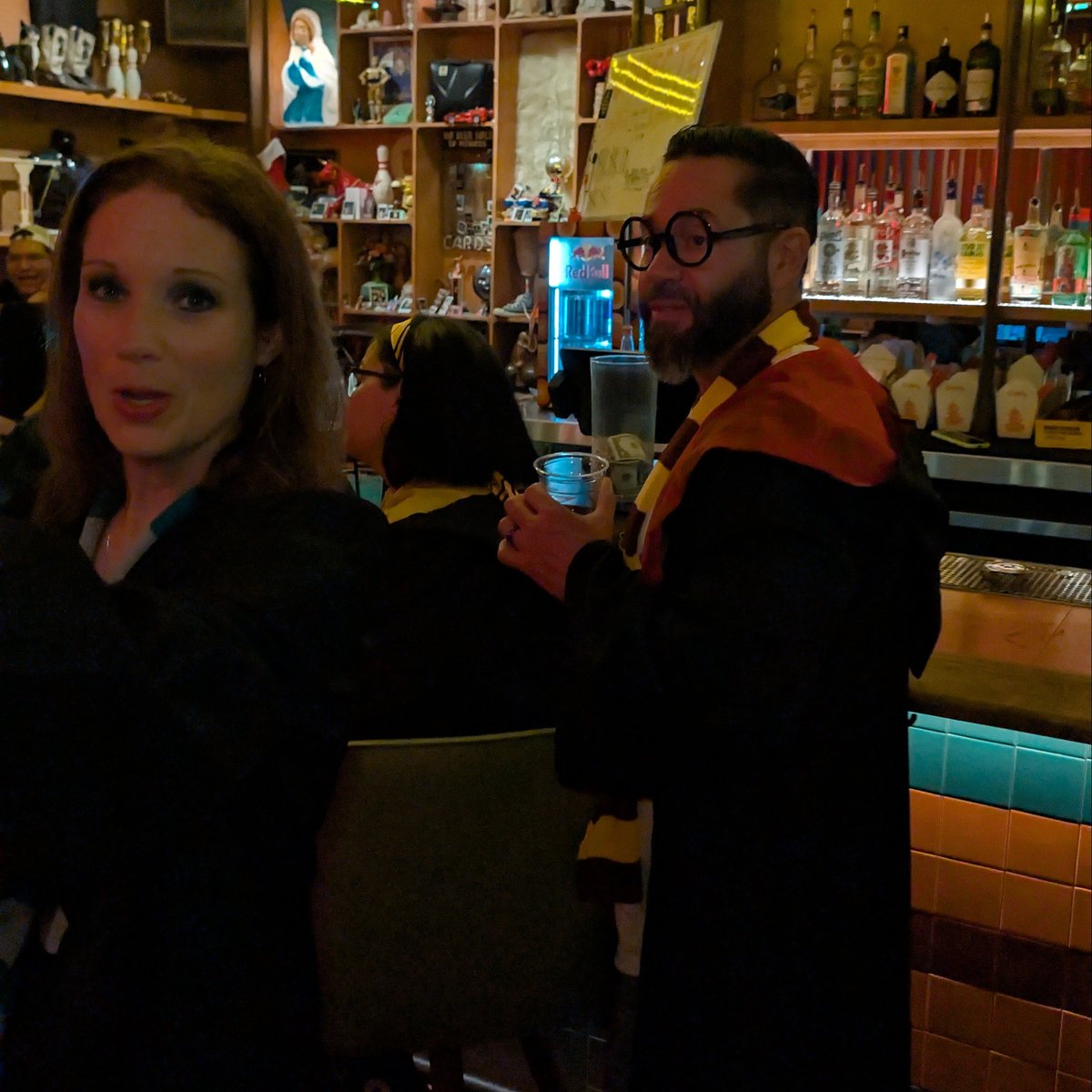 Spellbound couple in house scarves, robes, and round Potter glasses pause at a bar lined with potion-like bottles.
