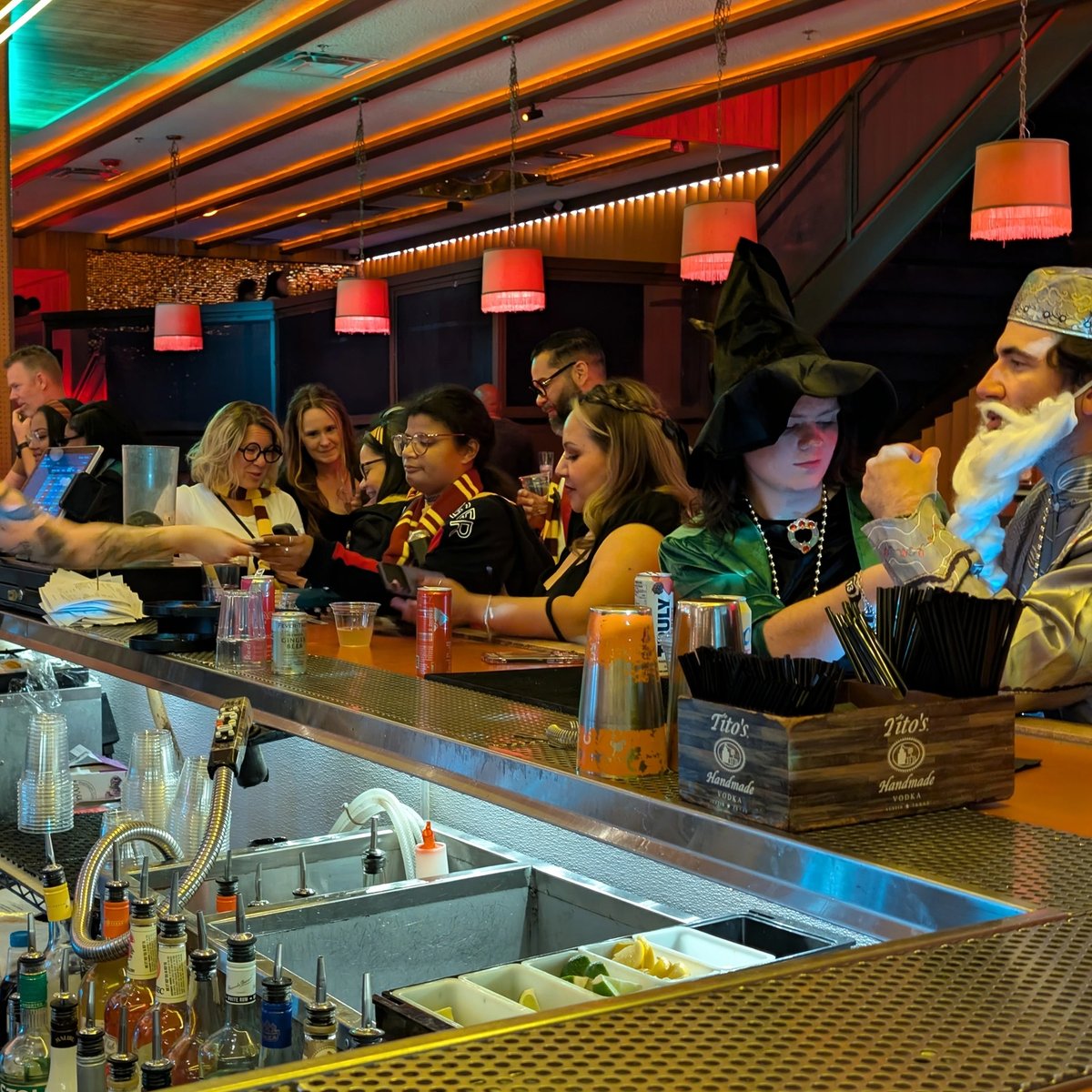 Witches and wizards crowd a wizard-crawl bar, a pointed-hat sorceress ordering a butterbeer-style cocktail.