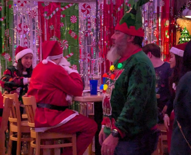 Full-suit Santa shares a table with a tiny Santa-hatted woman as an elf in a tall hat strolls past