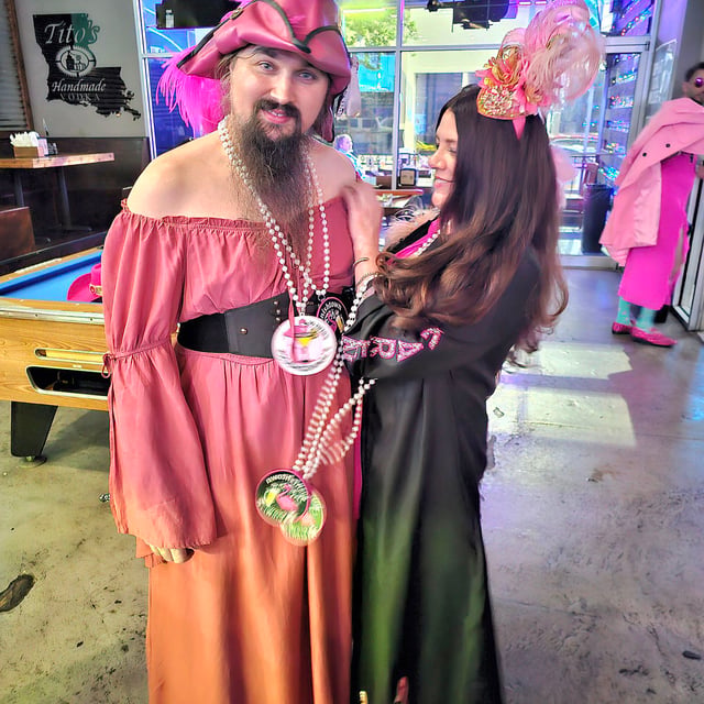 Bearded man in pink dress and tricorn hat poses with a woman in pink fascinator inside a pool bar
