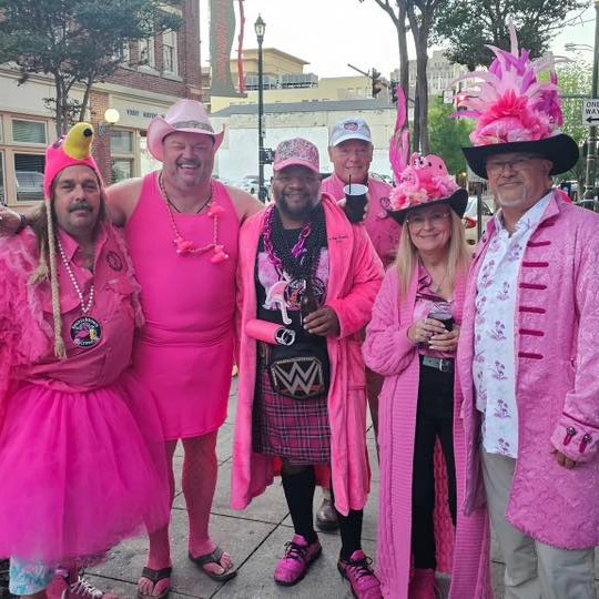 Six pink-dressed crawlers in feathered hats, tutus and wigs pose on a sidewalk under a streetlamp