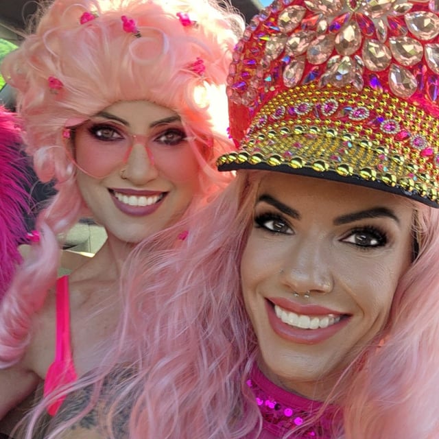 Tight selfie of two women in pink wigs, pink sunglasses and jeweled pink captain hat smiling