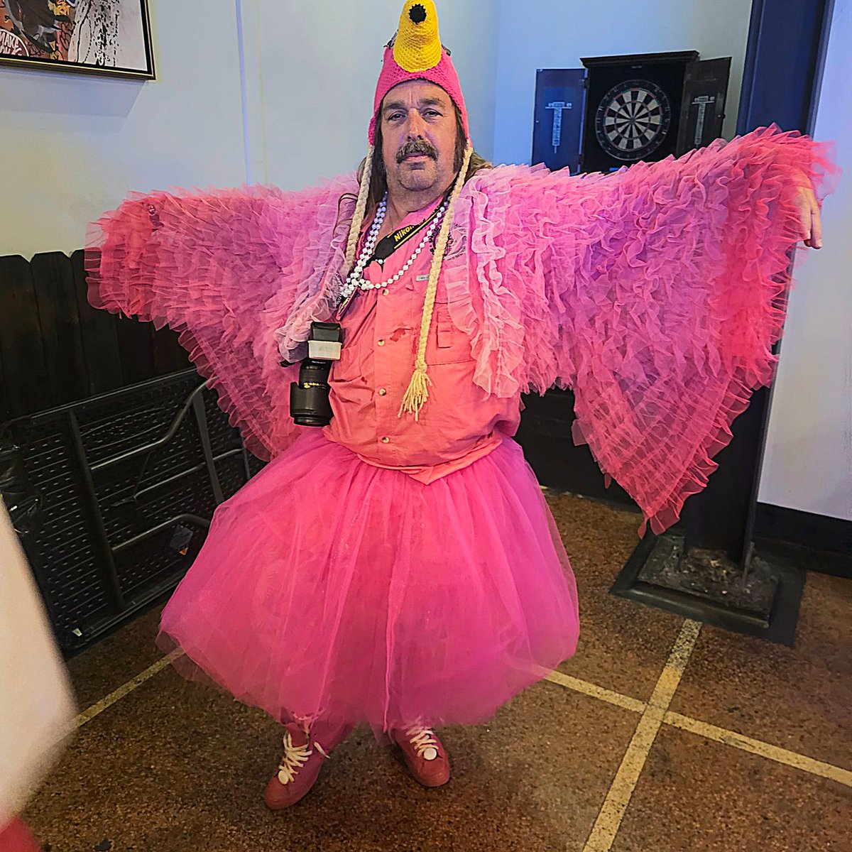Man in pink tutu, feathered wings and yellow knit hat poses with camera as a flamboyant pink bird
