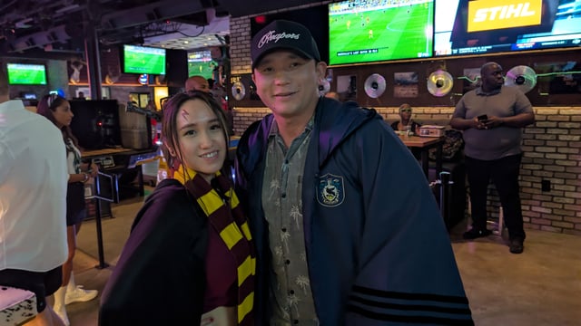 Lightning-scarred witch in house scarf cozies up to her robed wizard at a sports-bar stop on the wizard crawl.