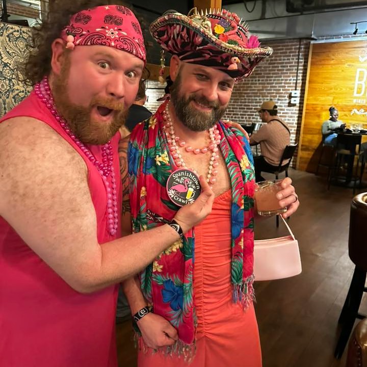 Two bearded men in pink dresses and beads pose inside a bar, one in a flowered hat holds a cocktail