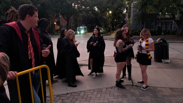 Robed wizard crawl crew checks phones streetside at dusk, house scarves and ties hanging loose between stops.