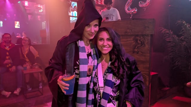 Two wizards-in-training in hooded Hogwarts-style robes and house scarves clutch a glowing blue potion at a Wizards Night Out stop.