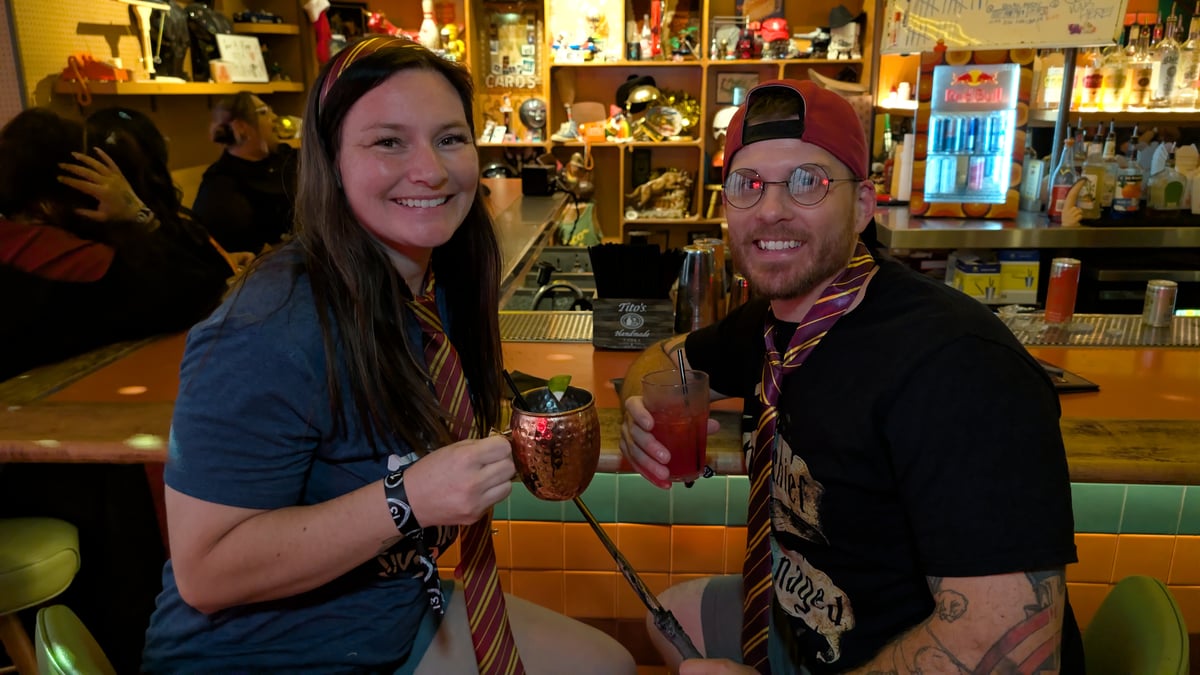 Couple in house ties toast with a copper mug and red potion, wand on the bar at a quirky Wizards Night Out stop.