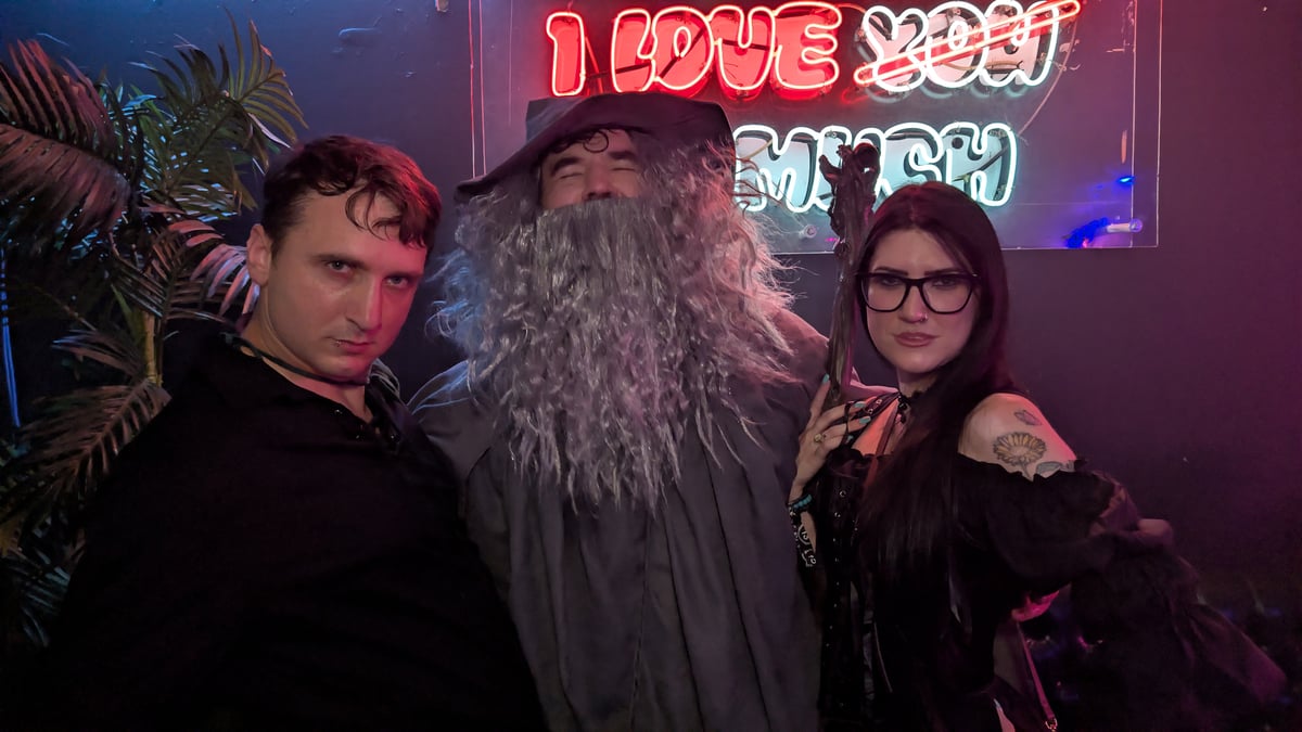 Long-grey-bearded wizard with pointed hat and staff flanks two attendees under neon on the wizard crawl