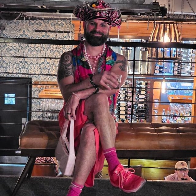 Bearded man in pink floral hat, dress and pink socks sits at a wine bar holding a small handbag