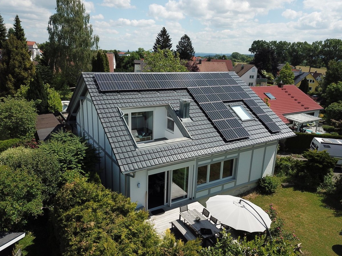 Aerial view of a detached house with a dark tiled pitched roof carrying photovoltaic modules around a dormer and a skylight, with a terrace and garden in front.