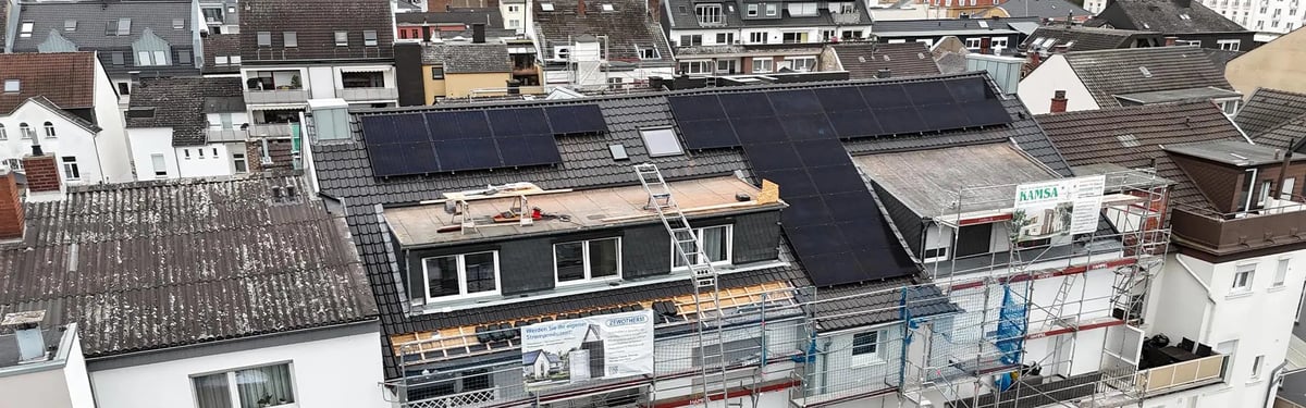 Aerial view of a row of multi-storey townhouses. Black photovoltaic modules sit on a dark pitched roof, in front of it a scaffolded top floor under renovation with a Zewotherm banner on the scaffolding.