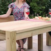 Solid wood outdoor dining table with a pink table runner, cheese, and wine glasses, set in a lush garden perfect for summer entertaining.