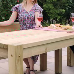 Solid wood outdoor dining table with a pink table runner, cheese, and wine glasses, set in a lush garden perfect for summer entertaining.