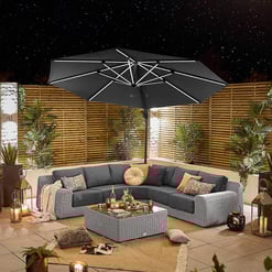 Stylish outdoor patio featuring a large black umbrella, cozy sectional sofa with assorted cushions, lush potted plants, decorative lanterns, and ambient lighting for relaxing evenings.