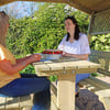 A woman in a yellow top and jeans receives a strawberry cake with strawberries from another woman in a white blouse seated at a wooden garden gazebo table. The scene is bright and surrounded by lush greenery.