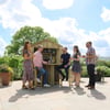 Outdoor garden party with a custom-built wooden bar, friends enjoying drinks, surrounded by lush greenery and potted plants on a sunny day.