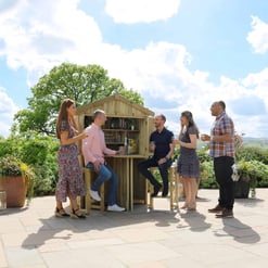 Outdoor garden party with a custom-built wooden bar, friends enjoying drinks, surrounded by lush greenery and potted plants on a sunny day.