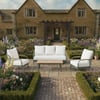 Lush garden patio with white outdoor furniture including a sofa and two chairs, surrounded by vibrant flowering plants and maintained hedges in front of a traditional stone cottage.