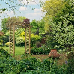 Lush garden with decorative wooden arbor, vibrant greenery, flowering plants, and outdoor terracotta pot, showcasing beautiful landscaping and garden design by Empire Home and Garden.