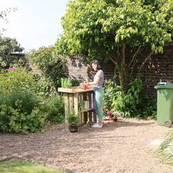 Lush garden outdoor space with a woman gardening at a wooden workbench, surrounded by vibrant plants, trees, and garden tools, showcasing the beauty of backyard landscaping and outdoor gardening.