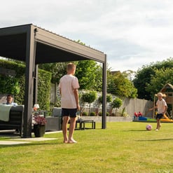 Modern backyard with a stylish pergola, children playing soccer, lush greenery, and outdoor furniture, showcasing garden ideas for entertaining and family fun in a beautiful outdoor space.