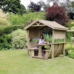 A woman sitting in a wooden garden gazebo surrounded by lush greenery and colorful flowering plants, ideal for outdoor relaxation and garden décor.