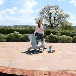 Adirondack-style garden chair on a patio with a woman standing nearby, enjoying a peaceful outdoor space with lush greenery and scenic rolling hills, perfect for outdoor living and relaxation.