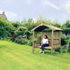 Rustic wooden garden shelter with a woman sitting on a bench in a lush backyard, surrounded by vibrant flowers, trees, and well-maintained lawn, ideal for outdoor relaxation and garden enhancement.