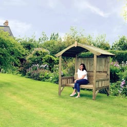 Rustic wooden garden shelter with a woman sitting on a bench in a lush backyard, surrounded by vibrant flowers, trees, and well-maintained lawn, ideal for outdoor relaxation and garden enhancement.