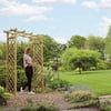 Relaxing garden scene with a woman assembling a wooden arbor in a lush, green backyard with vibrant trees and blooming flowers, perfect for outdoor living and garden decoration.