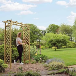 Relaxing garden scene with a woman assembling a wooden arbor in a lush, green backyard with vibrant trees and blooming flowers, perfect for outdoor living and garden decoration.