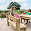 Beautiful outdoor dining scene with two women enjoying a meal on a wooden patio table surrounded by lush garden in summer. Perfect for garden furniture, outdoor dining, and patio decor inspiration.