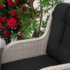 Luxurious outdoor wicker armchair with dark cushions, surrounded by vibrant red flowering plants and lush green foliage, perfect for relaxing garden moments.