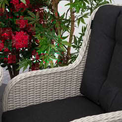 Luxurious outdoor wicker armchair with dark cushions, surrounded by vibrant red flowering plants and lush green foliage, perfect for relaxing garden moments.