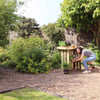 Lush backyard garden with a woman tending to potted plants near a brick wall, featuring garden furniture, vibrant greenery, and floral accents, perfect for outdoor gardening and relaxation.