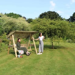 Picture of two women enjoying a sunny day on a wooden garden swing with lush greenery and blooming flowers, perfect for outdoor relaxation and garden furniture.