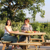 Girls enjoying outdoor dining at a wooden picnic table surrounded by lush garden greenery with a large tree in the background.