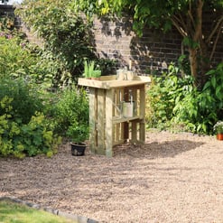 A backyard garden with a wooden outdoor sink station, surrounded by lush green plants, flowering bushes, and a brick wall, ideal for outdoor kitchen and garden maintenance.