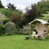 Garden with a wooden garden bench with canopy and colorful cushions, surrounded by lush flowering plants and trees, ideal for outdoor relaxation and garden décor.