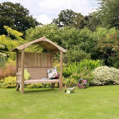 Cozy wooden garden bench with canopy surrounded by lush greenery and colorful flowering plants in a landscaped yard.