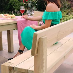 Warm light wood outdoor dining table and bench set with a pink checkered table runner, enjoying a summer garden meal with a cheese platter and glasses of rosé wine.
