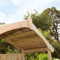 Beautiful wooden arbor with curved roof in a lush garden setting featuring colorful flowers and tall trees, perfect for outdoor relaxation and garden enhancement.