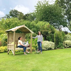 A peaceful garden scene with two women enjoying outdoor relaxation, surrounded by lush greenery, flowering plants, and a wooden garden bench under a small pergola. Ideal for outdoor living and garden decor.