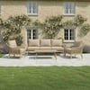 Stylish outdoor patio furniture set with beige cushions, surrounded by lush green lawn and flower beds in front of a charming brick house with blooming climbing roses.